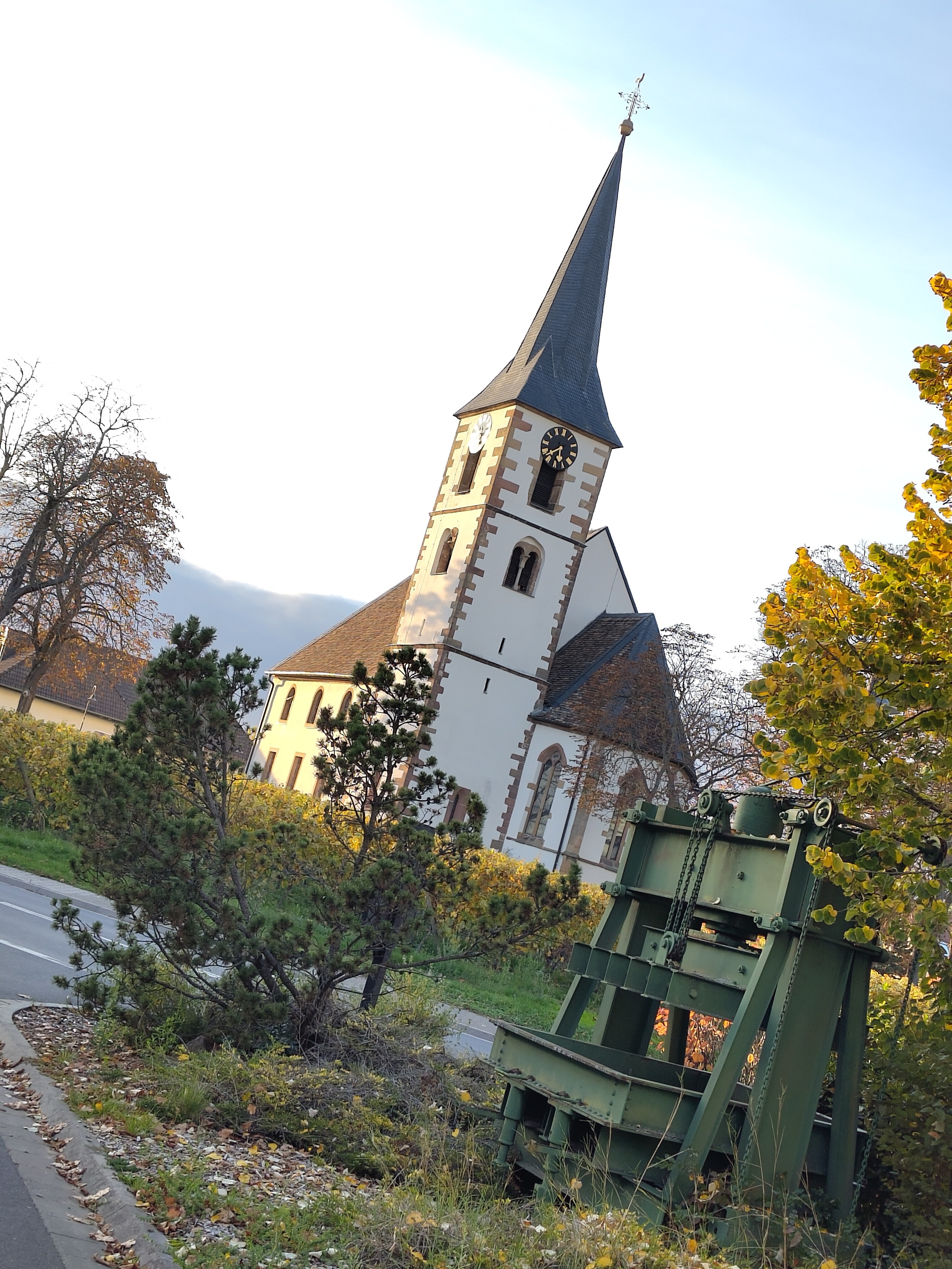 Foto einer kleinen Kirche mit spitzem Turm im Abendlicht, schräg im Dutch Angle aufgenommen. Im Vordergrund eine buschige kleine Kiefer und rechts daneben, halb unter einem Baum, eine Weinkelter aus Stahlprofilen. 